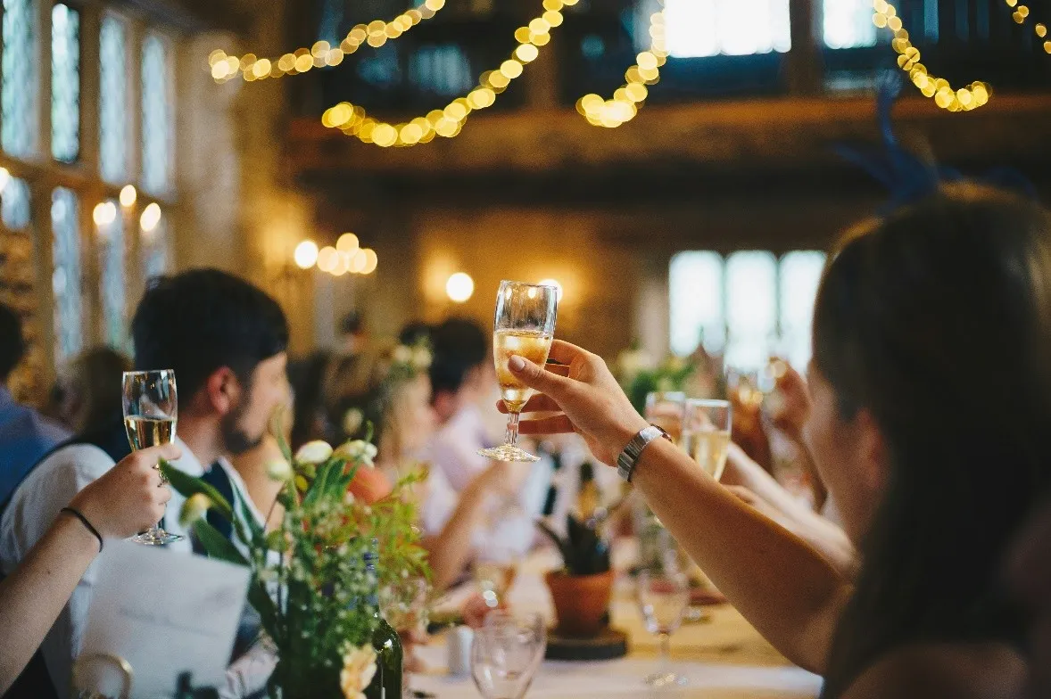 Detail shot of a hand raising a champagne glass during a toast at a wedding banquet. The scene takes place in an elegant rustic hall with large windows. In the blurred background, other guests are seen toasting at tables decorated with floral arrangements under strings of warm lights that create a festive and cozy atmosphere.