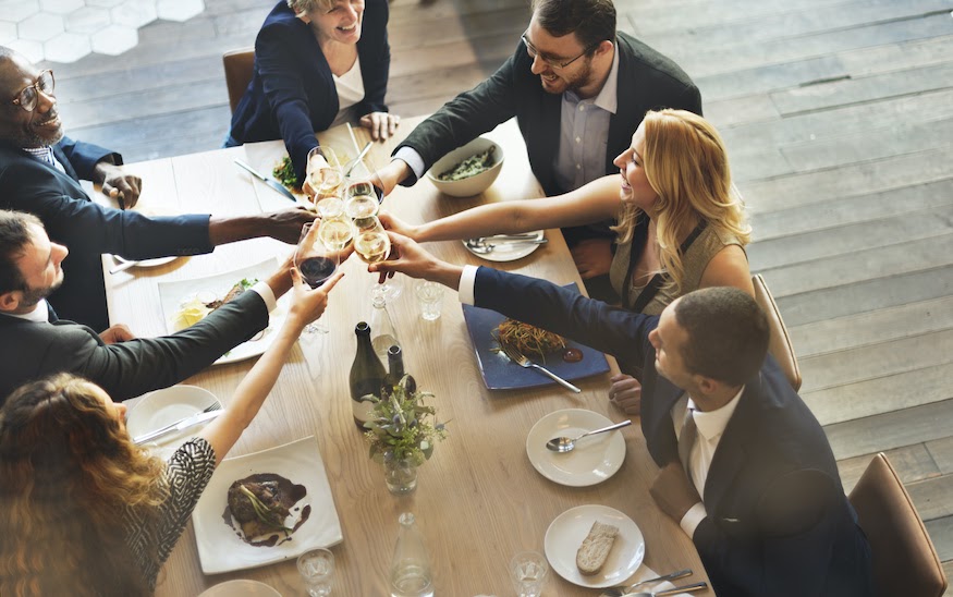 High-angle view of a diverse group of professionals sitting at a wooden table during a business lunch. Everyone is smiling and toasting with glasses of white and red wine in the center of the table. Gourmet dishes with meat and sides, wine bottles, and simple floral decor are visible in a modern office setting with wooden floors.