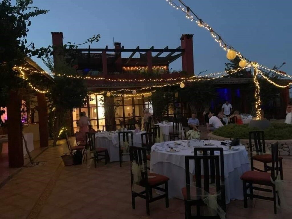 An outdoor terrace at night with round tables set for a wedding banquet, illuminated by warm string lights hanging over the courtyard of a rustic estate.