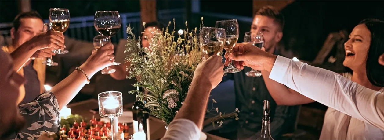 Medium shot of a nighttime toast during a private celebration. Several hands are shown raising glasses of white and red wine in a festive and joyful atmosphere. In the center of the table, a wild floral arrangement and lit candles provide warm lighting. On the right, a woman laughs openly while participating in the toast with her friends.