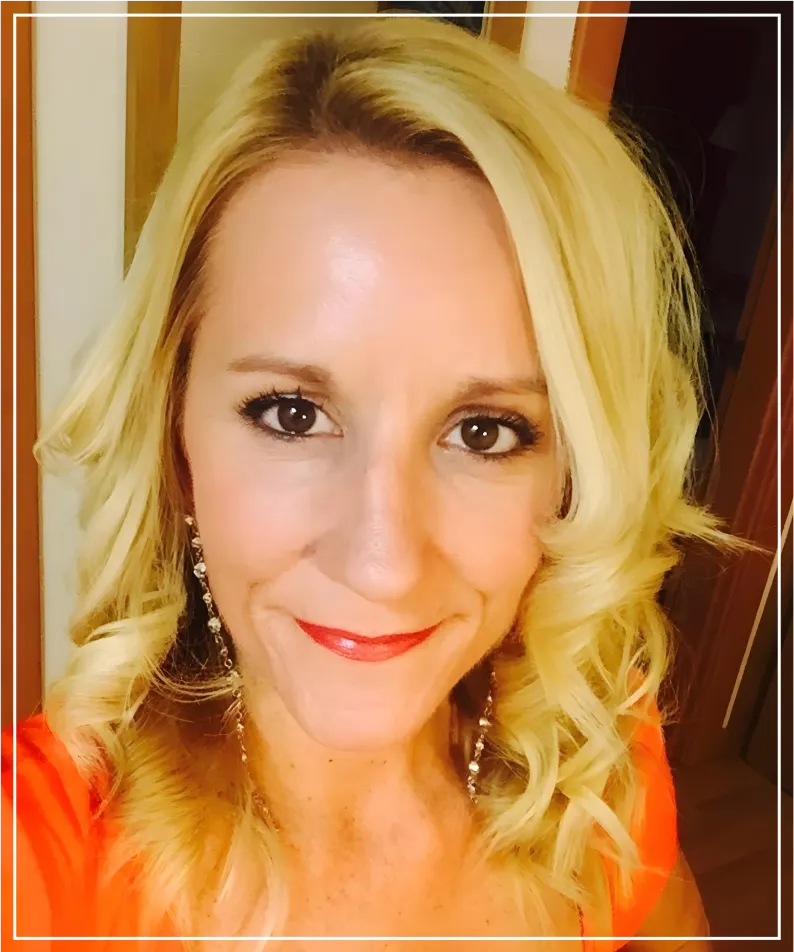 Close-up portrait of a blonde woman with wavy hair. She is wearing a bright orange garment and looks directly at the camera with a friendly smile. The background is neutral, and the lighting highlights her features clearly and professionally.