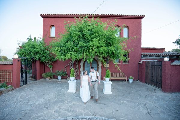 An elegant newlywed couple poses serenely under the shade of a large, leafy tree that stands at the estate's entrance. The scene is characterized by the striking contrast between the bride's pure white dress and the vibrant clay-red facade of the main building, while classic white planters and a stone path complete this rustic and stately corner.