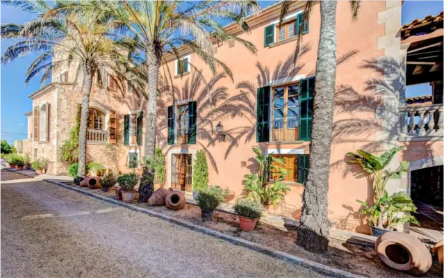 Facade of a terracotta-colored Mediterranean villa with green shutters, palm trees casting shadows on the wall, and large decorative clay pots.