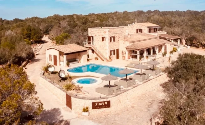 Aerial view of a large rural estate featuring a two-story stone house, a crystal-clear blue pool, and extensive surrounding green areas.