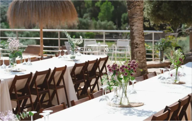 Outdoor event setup with long tables dressed in white linens and flowers, located on a terrace with a metal railing under the shade.