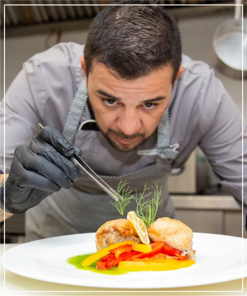 Photograph of a professional chef concentrated while plating a gourmet dish. He is wearing a gray apron and black kitchen gloves. Using precision tweezers, he carefully places a detail on a serving of meat decorated with colorful peppers on a white plate.