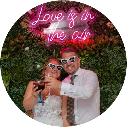 Circular photograph of a couple celebrating in a festive setting. Both are wearing white heart-shaped sunglasses and holding cocktail glasses. In the background, a green foliage wall stands out with a pink neon sign that says "Love is in the air."
