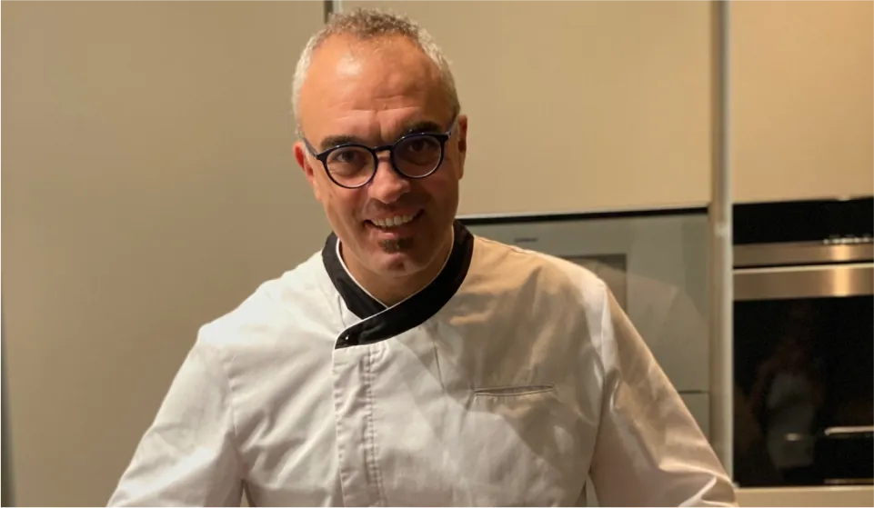 Frontal portrait of Sergio Rifugio, Director of Mallorca Weddings And Events, smiling, wearing glasses and a white culinary jacket with black trim, posing in a brightly lit professional kitchen.