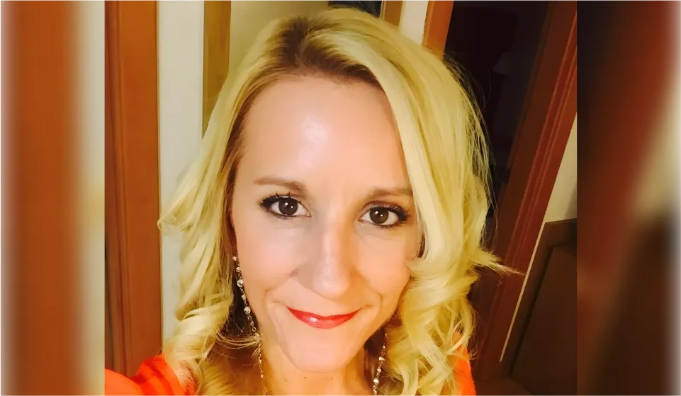 Close-up of a blonde woman smiling at the camera, wearing elegant earrings, natural makeup with red lips, and an orange dress.