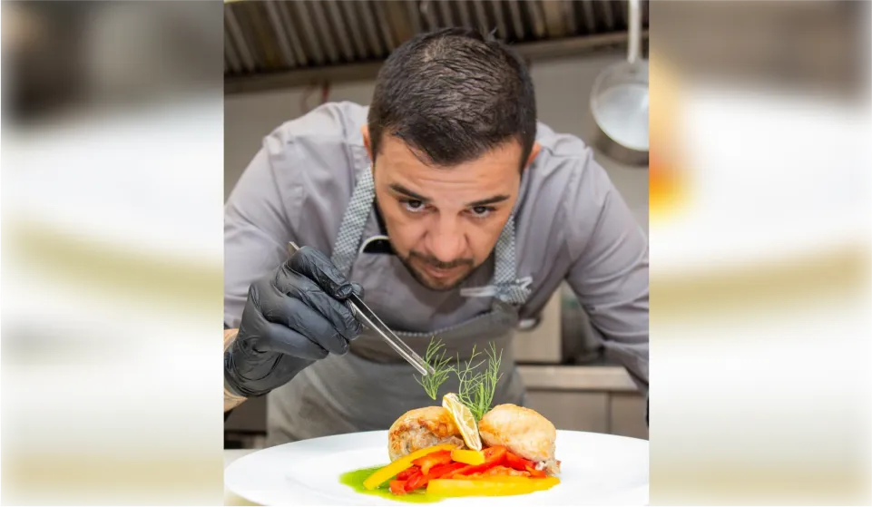 Close-up of a chef wearing black gloves using precision tweezers to delicately place fresh herbs onto a plate of sautéed vegetables.