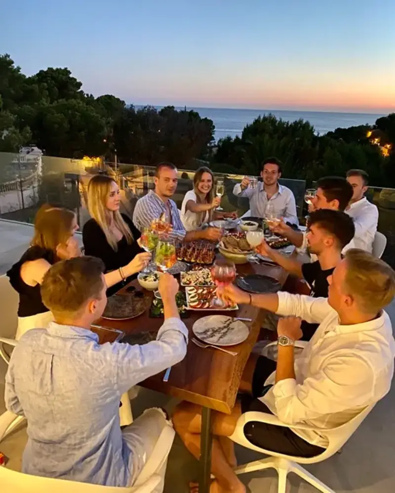 A group of friends dines outdoors on a terrace during sunset, with direct views of the Mediterranean Sea on the horizon. They are seated around a rustic wooden table filled with plates, food platters, and wine glasses. The diners are cheerfully toasting with their glasses raised under warm ambient light while the sky shows gradients of blue and orange.