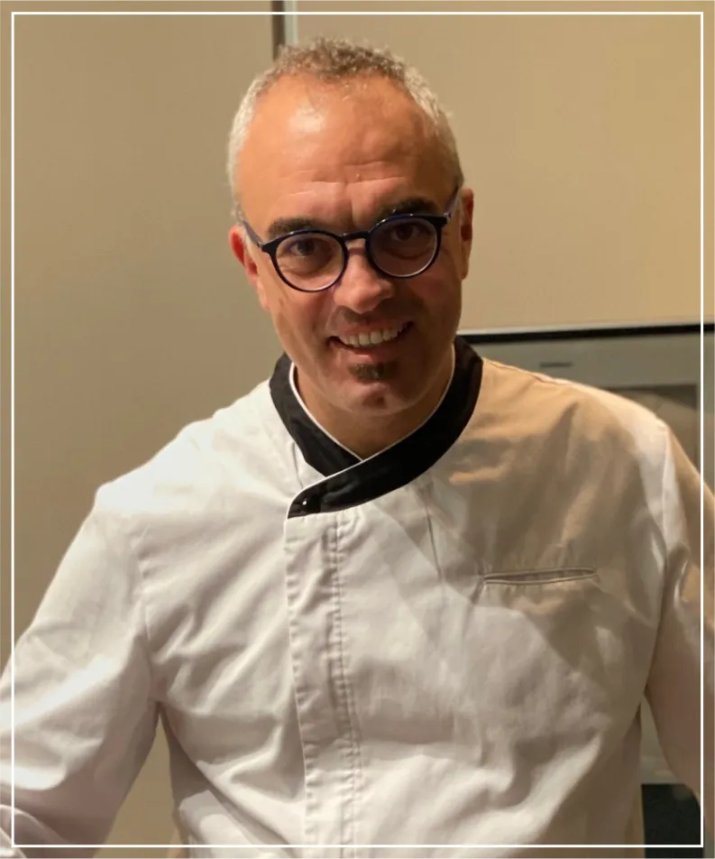 Frontal portrait of Sergio Rifugio, Director of Mallorca Weddings And Events, smiling, wearing glasses and a white culinary jacket with black trim, posing in a brightly lit professional kitchen.