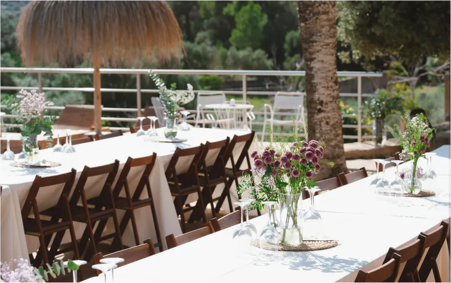 Outdoor event setup with long tables dressed in white linens and flowers, located on a terrace with a metal railing under the shade.