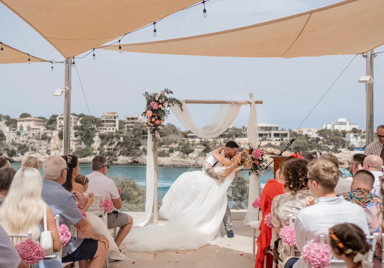 Outdoor wedding ceremony held under a large canopy with a floral arch, guests seated, and a beautiful sea view in the background.